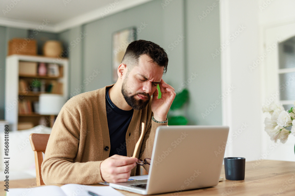 Young Caucasian Man in Pain Working from Home at Desk with Laptop in Bright, Well-Lit Room Young Caucasian Man in Pain Working from Home at Desk with Laptop in Bright, Well-Lit Room