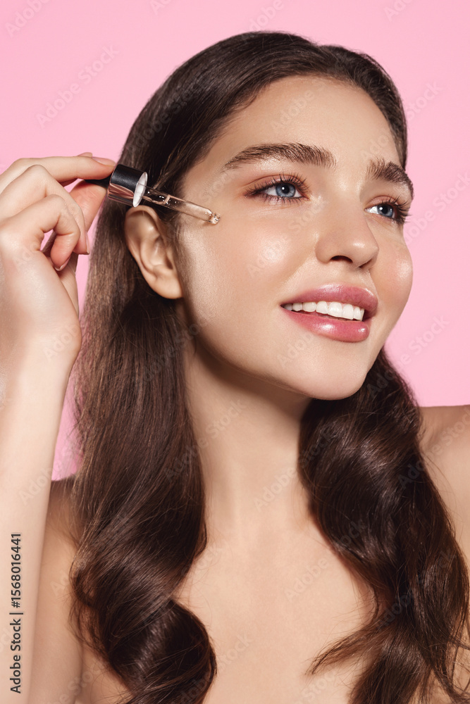 Vertical portrait of beautiful beauty model, holds a dropper near cheek, applies skin care moisturiser, hydrating serum with hyaluronic acid and collagen, pink background Vertical portrait of beautiful beauty model, holds a dropper near cheek, applies skin care moisturiser, hydrating serum with hyaluronic acid and collagen, pink background