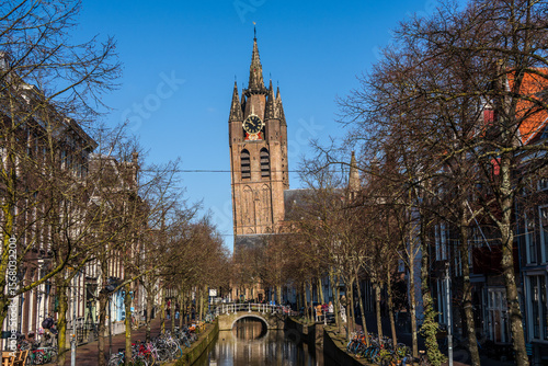 Bright winter morning in Delft. The brick tower of the Nieuwe Kerk stands at the canal’s end, its clock and spires framed by leaf-bare trees, parked bicycles, and narrow Dutch houses. 