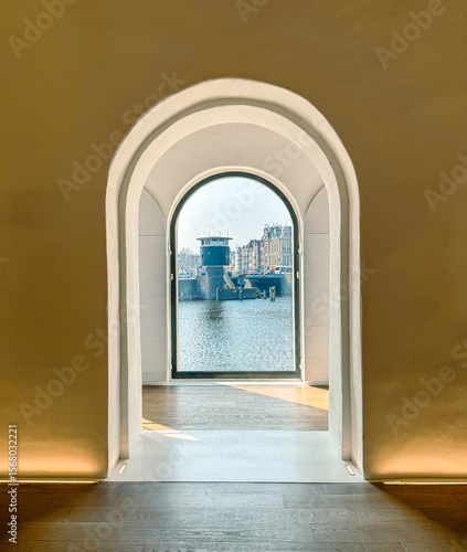 Inside the National Maritime Museum, warm-lit stucco walls and a clean arched doorway create a minimalist frame. The window looks onto the Oosterdok basin, where a small bridge control tower and class
