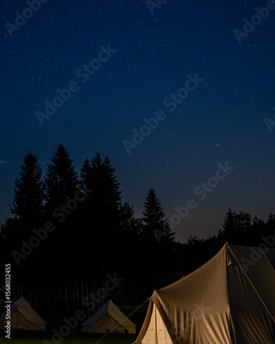 Glamping bell tent glows warmly in a dark clearing while the night sky stretches overhead. Good for camping or outdoor-life decor.
