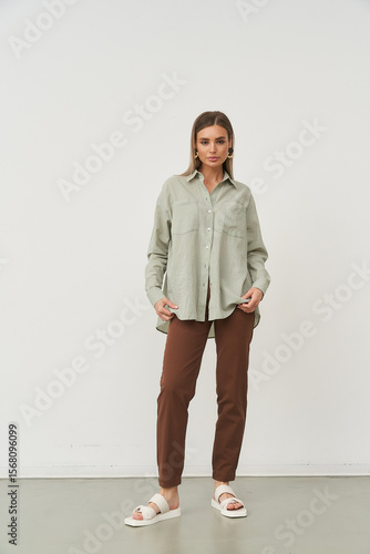 Neutral expression, woman stands confidently wearing a light gray button-up shirt and brown trousers with white sandals against a plain backdrop. She holds the shirt lightly with both hands