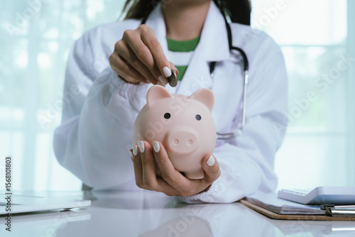 Close up young female general practitioner putting coin in piggy bank, managing hospital budget using laptop accounting applications, medical insurance finances, healthcare money savings concept.