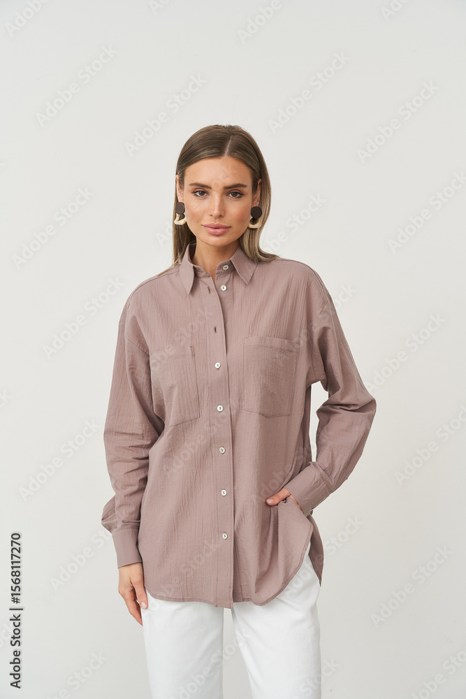 Woman wearing oversized mauve button-up shirt and white pants with hand in pocket. She has straight brown hair, gold hoop earrings, and a neutral expression. Studio lighting Woman wearing oversized mauve button-up shirt and white pants with hand in pocket. She has straight brown hair, gold hoop earrings, and a neutral expression. Studio lighting