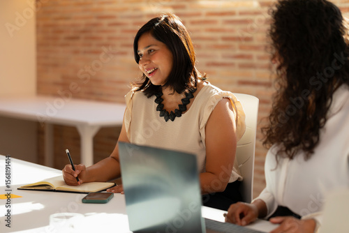 A young woman, with Indian Asian features, smiles while chatting with her teammates during a meeting in the office in the office