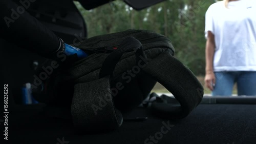 Close-up of gray backpack with tennis racket lies in closed trunk, door opens and sporty woman in white t-shirt takes bag and leaves. Sporty lifestyle and using car for training. No face