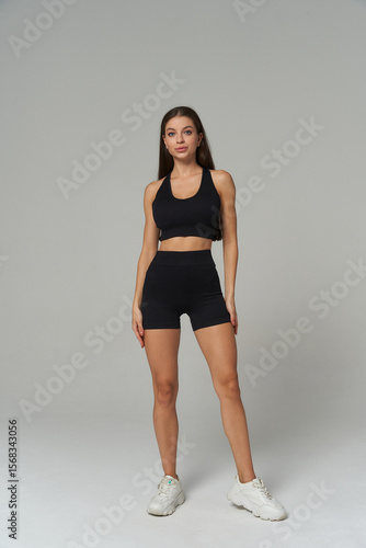 Athletic woman standing confidently in matching black sports bra and shorts, paired with white sneakers. Her long hair is down, and she maintains a neutral expression facing forward