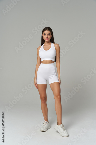 A woman stands confidently in white athletic wear, consisting of a sleeveless crop top and high-waisted shorts. She pairs the outfit with white sneakers, posing against a neutral backdrop