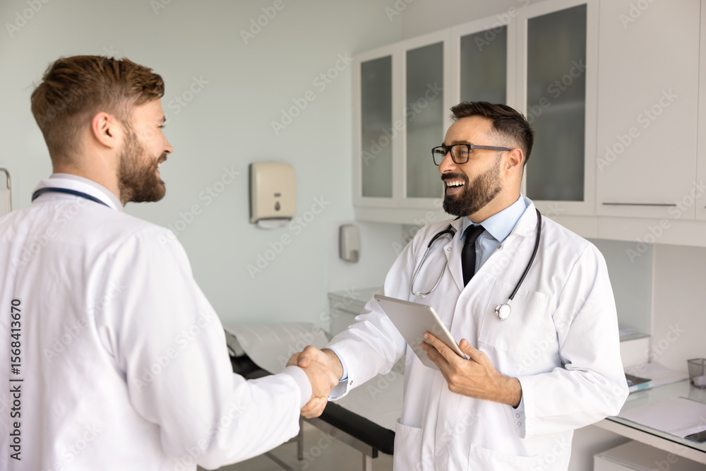 Two positive medical practitioners shaking hands in hospital exam room, smiling, laughing, successfully working together. Latin doctor giving handshake to colleague, thanking for help, teamwork Two positive medical practitioners shaking hands in hospital exam room, smiling, laughing, successfully working together. Latin doctor giving handshake to colleague, thanking for help, teamwork