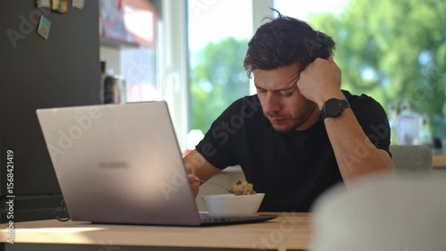 Man sitting at kitchen table, eats oatmeal porridge with displeased look and pushed her plate away. Unhappy man eating tasteless breakfast in kitchen table with laptop
