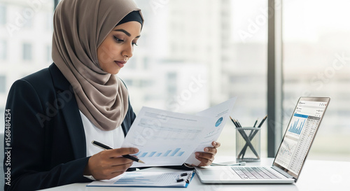 Executive Indian or Arab businesswoman in hijab working at desk with documents and laptop, analyzing financial data in a modern office environment.