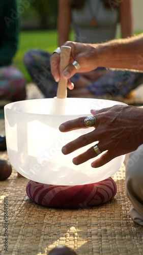 Close-up video of hands playing white crystal singing bowls with mallets on outdoor wooden surface, soft-focus background shows people on mats, highlighting ritual of sound healing and relaxation
