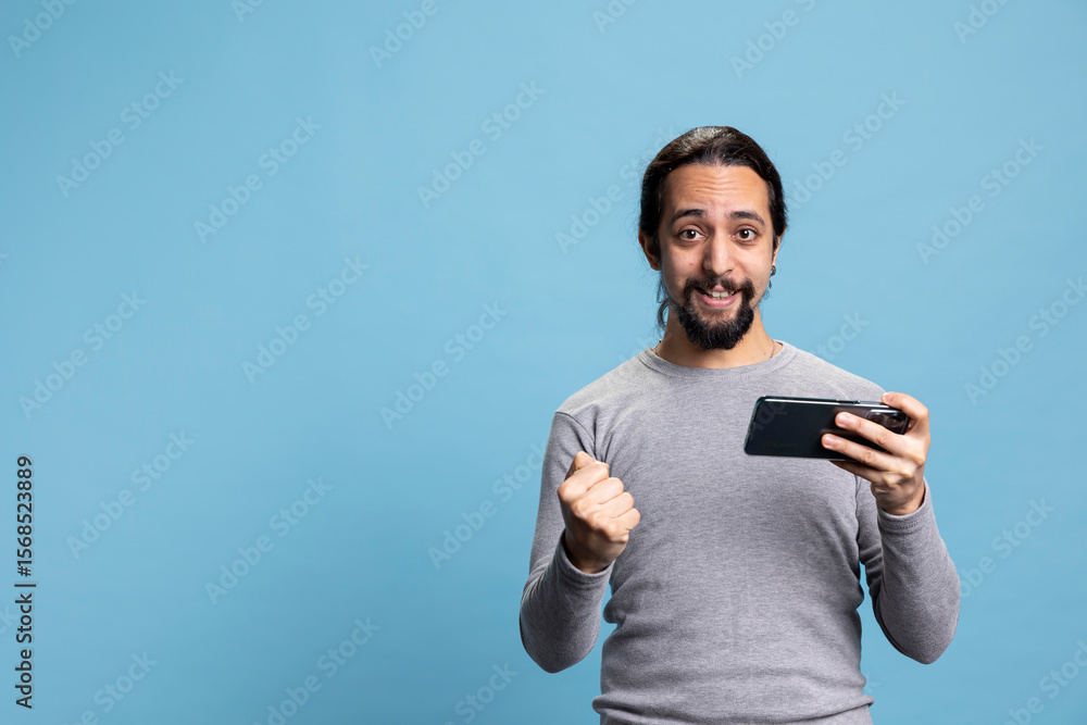Overjoyed young man winning an online tournament in front of camera, feeling pleased and proud of his win against blue background. Satisfied person smiling in studio, plays video games. Overjoyed young man winning an online tournament in front of camera, feeling pleased and proud of his win against blue background. Satisfied person smiling in studio, plays video games.