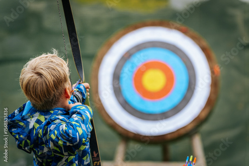 A young boy aims a bow at a colorful archery target, focusing intently while holding the string back, wearing a vibrant blue patterned jacket.