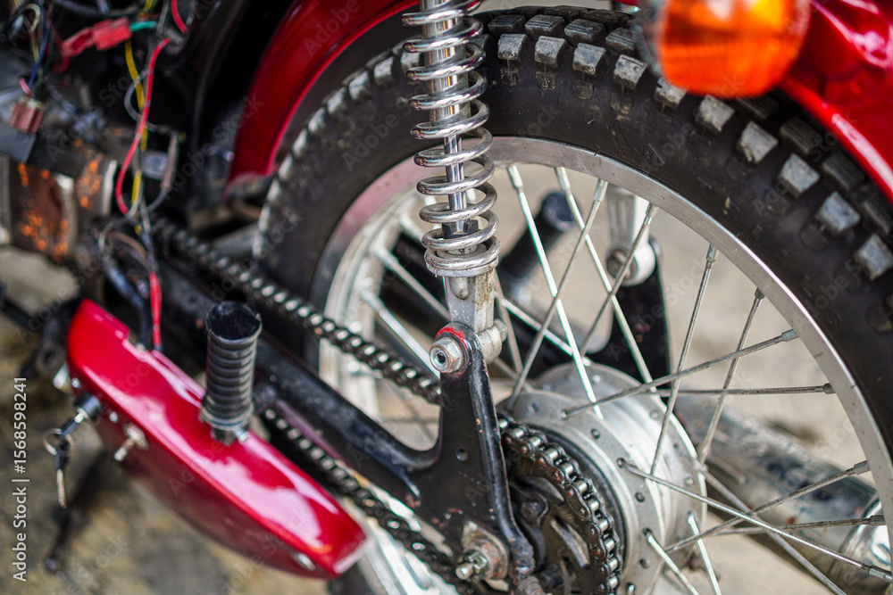 Obraz premium A close-up shot showcases the rear wheel assembly of a vintage motorcycle. The silver suspension spring, tire, chain, and red fender create a visually striking composition with depth.