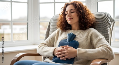 Young woman in cream sweater and blue jeans embracing a hot water bottle while relaxing in a chair near a window with natural light. Focus on comfort and well-being.