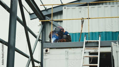 A construction worker performs welding on a steel structure at a high elevation, wearing proper protective gear including a welding helmet, gloves, and safety harness
