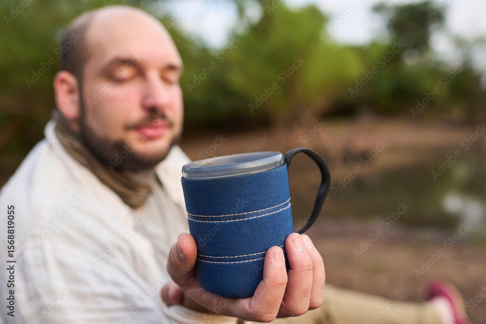 Man offers towards the camera a cup of freshly brewed coffee in nature, early in the morning. His expression is one of tranquility, calm and well-being. El Palmar National Park, Entre Rios, Argentina. Man offers towards the camera a cup of freshly brewed coffee in nature, early in the morning. His expression is one of tranquility, calm and well-being. El Palmar National Park, Entre Rios, Argentina.