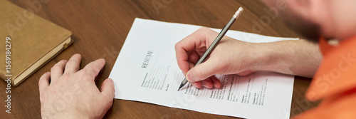 Caucasian young adult man filling out resume form on wooden desk, hand holding pencil, notebook nearby, participating in educational program focused on rehabilitation in prison setting