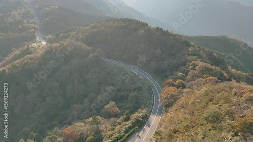 Aerial forward drone shot over a winding mountain road with scattered cars. The landscape features dense green and some autumnal trees. Distant mountains under a clear blue sky are visible during day