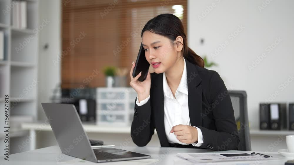 Asian businesswoman sitting at her desk in a modern office, talking on the phone while analyzing data on her laptop and pointing at the screen Asian businesswoman sitting at her desk in a modern office, talking on the phone while analyzing data on her laptop and pointing at the screen