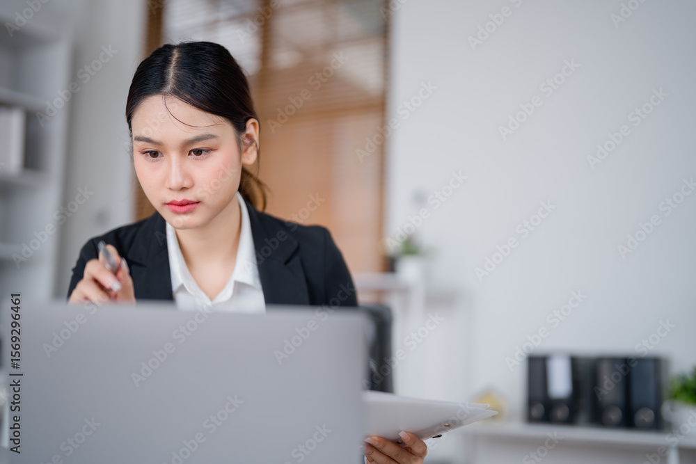 Young Asian businesswoman holding a pen and reading financial documents while working on a laptop at an office desk, focused on project strategy and report preparation Young Asian businesswoman holding a pen and reading financial documents while working on a laptop at an office desk, focused on project strategy and report preparation