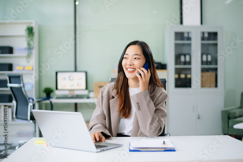 Confident young Asian businesswoman smiles while multitasking talking on her smartphone and using her laptop at a modern office desk
