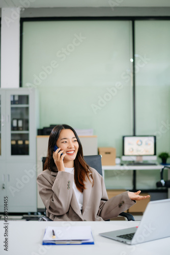 Confident young Asian businesswoman smiles while multitasking talking on her smartphone and using her laptop at a modern office desk