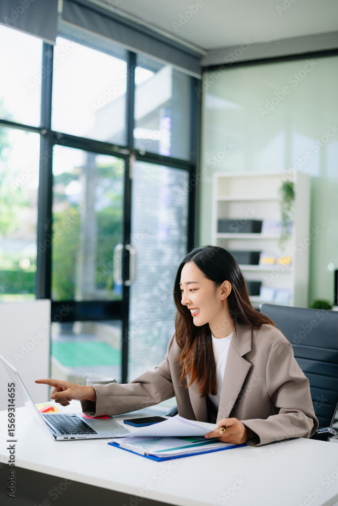 Smiling young Asian businesswoman points at her laptop while reviewing documents at a modern office desk Smiling young Asian businesswoman points at her laptop while reviewing documents at a modern office desk