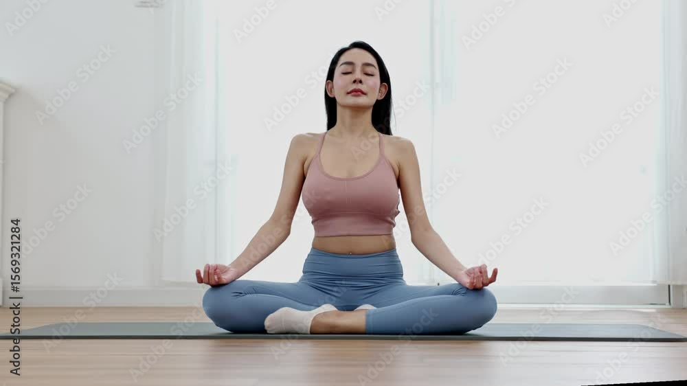 Asian Young woman practicing meditation in lotus pose on yoga mat indoors wearing comfortable activewear, expressing calm and focus with natural light enhancing fitness and wellness atmosphere Asian Young woman practicing meditation in lotus pose on yoga mat indoors wearing comfortable activewear, expressing calm and focus with natural light enhancing fitness and wellness atmosphere