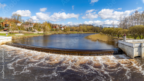 Pskov Dam on Pskova River with turbulent water flow over concrete spillway, surrounded by natural grassy banks and spring greenery. Sunny day in Pskov, Russia. 