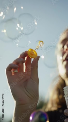 Human hand blows iridescent soap bubbles, macro video capturing vibrant rainbow colors. Shimmering spheres float against a soft, sun-drenched background with bokeh. Slow-motion low-angle video in
