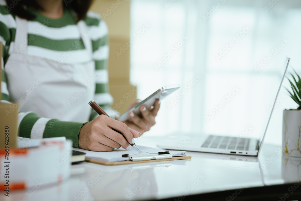 Fototapeta premium An Asian female entrepreneur works on her laptop to manage the logistics of her e-commerce business, organizing parcels, processing orders, and managing online sales and deliveries from her home offic