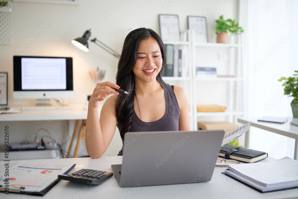 Smiling young woman working on a laptop at her desk, surrounded by charts, reports, and office supplies. Smiling young woman working on a laptop at her desk, surrounded by charts, reports, and office supplies.