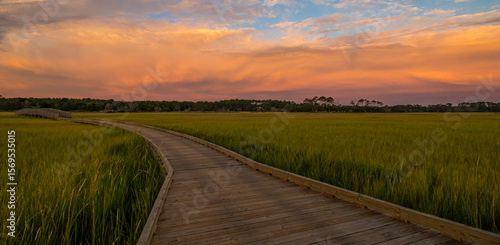 A boardwalk crosses over a salt marsh at sunrise with the clouds reflecting the light from the morning sun
