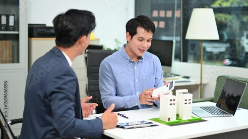 Wallpaper Mural Two real people in business attire collaborate over documents and property models at a modern office desk, illustrating teamwork, real estate planning Torontodigital.ca