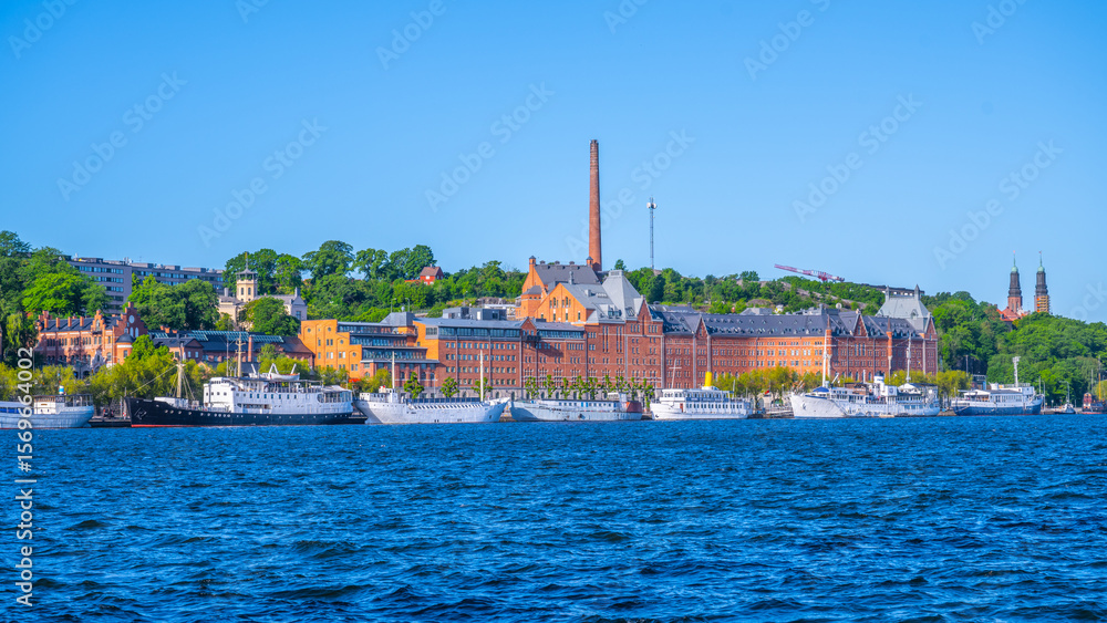 Obraz premium A vibrant view of Stockholm shows historic buildings along the waterfront. Ships line the harbor against a clear blue sky, highlighting the beauty of the city during a sunny day.