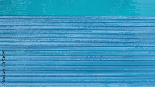 Swimming pool covered with an automatic shutter, water droplets on the surface. Top-down view, the panel texture forms a rhythmic pattern in cool blue tones
