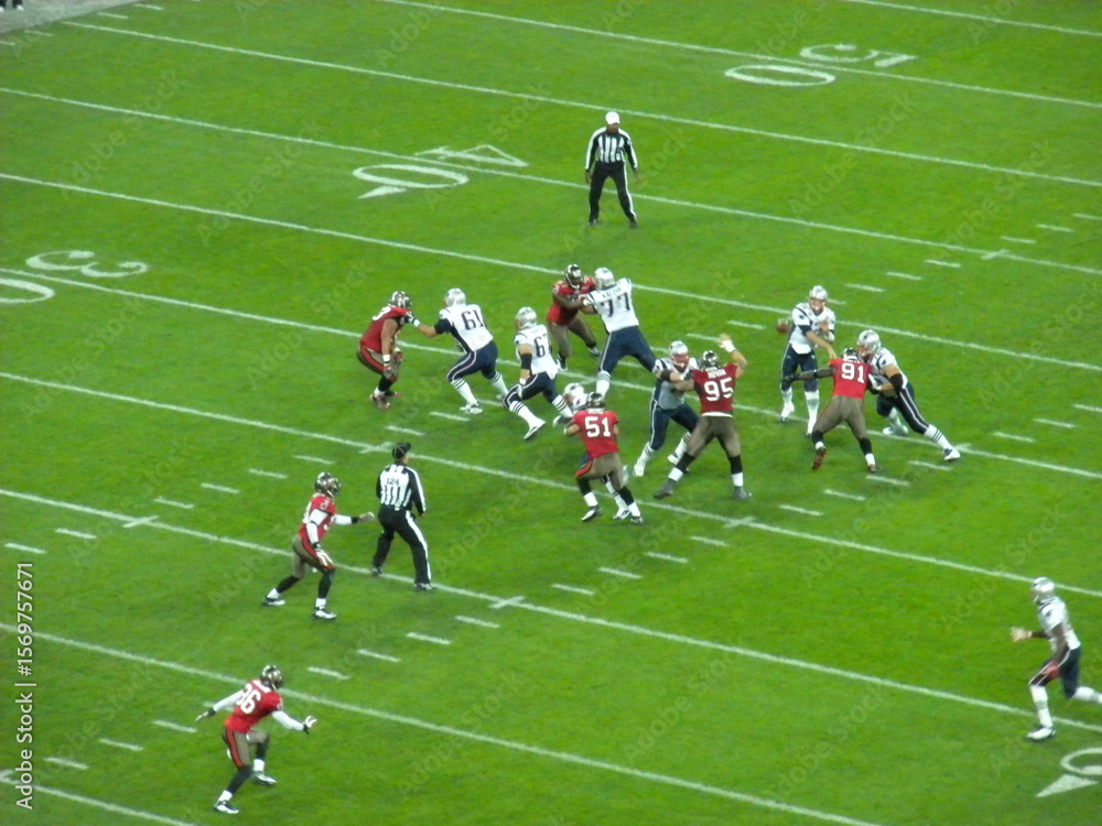 Fototapeta premium American football game in action between two teams on a grassy stadium field, with players in red and white uniforms and referees observing play