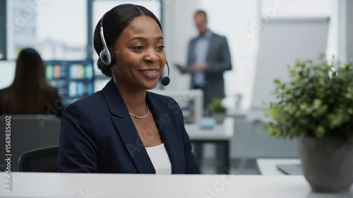 A professional woman wearing a headset smiles while assisting a customer at her desk in a modern office, with a blurred colleague in the background engaged in a discussion