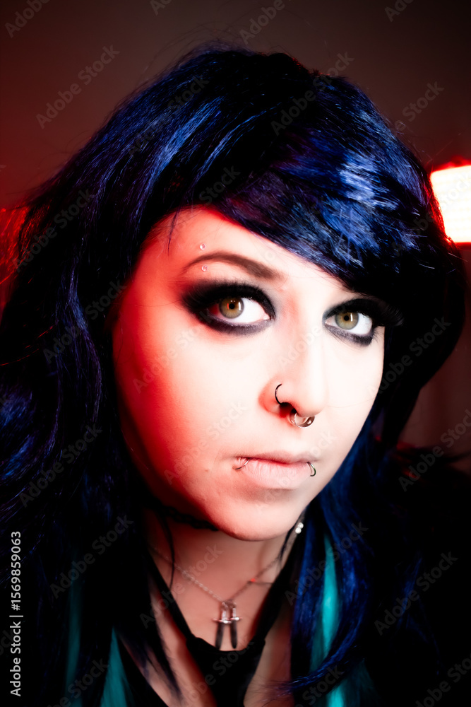 Alternative girl with dark makeup, striped sleeves, and facial piercings posing indoors under moody red and purple lighting. Alternative girl with dark makeup, striped sleeves, and facial piercings posing indoors under moody red and purple lighting.