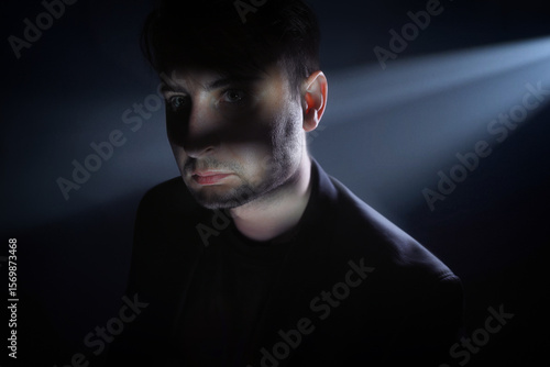 Dramatic close-up portrait of a young man with intense green eyes, partially hidden in shadow. A sharp beam of light cuts across his face, creating a mysterious and cinematic atmosphere. The man wears