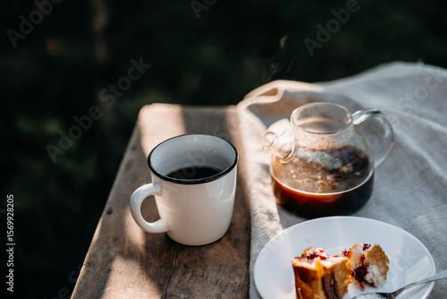 A white cup of hot coffee, a glass coffee pot, and a cherry pie on a plate rest on a wooden table glowing with gentle morning backlight, evoking a peaceful cottagecore mood.