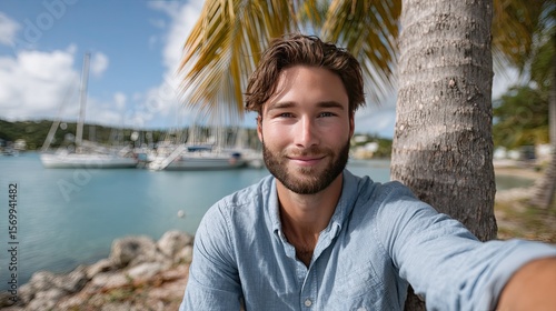 A young man takes a selfie on a beach with sailboats and palm trees behind him, capturing a sunny day
