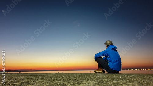 Lone man in a blue jacket sits on a pier, contemplating serene scene as the last light of sunset fades into a deep blue night sky over the water, capturing a moment of quiet introspection and peace.