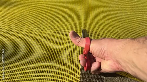 Close-up footage of a person cutting yellow fiberglass facade mesh with red-handled scissors on a rough concrete surface in bright sunlight, showing strong shadows and texture detail.
