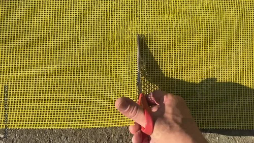 Close-up footage of a person cutting yellow fiberglass facade mesh with red-handled scissors on a rough concrete surface in bright sunlight, showing strong shadows and texture detail.