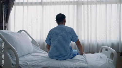 Rear view of sick old man sitting on bed looking at window inside hospital in lonely, depressed and hopeless attitude, health concept.