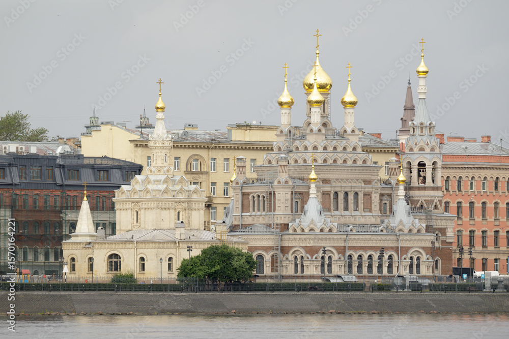 Naklejka premium Skorbyashchenskaya Church in Saint Petersburg, featuring ornate architecture and golden onion domes, located by the riverside against historic buildings.