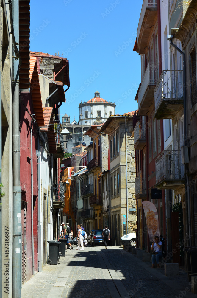 Fototapeta premium Narrow cobblestone street in Porto, Portugal, lined with colorful historic buildings and crowned by the iconic Serra do Pilar Monastery under a clear blue sky